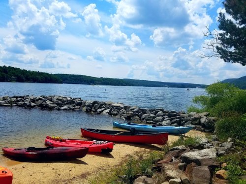Colorful kayaks on a sandy shore by a calm lake, with rocky outcrops and a blue sky filled with fluffy clouds.