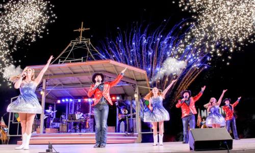 A lively performance on stage with singers in sparkly outfits, fireworks lighting up the night sky behind them.