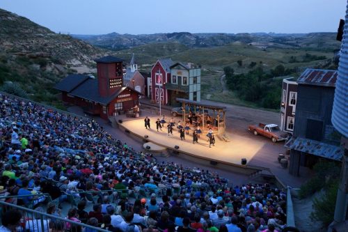 A large audience watches a performance on an outdoor stage surrounded by scenic hills and colorful buildings.