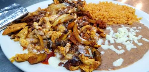 A plate of chicken fajitas with onions, rice, refried beans, and cheese, served with a silver fork on the side.