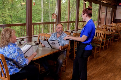 A waitress takes an order from a couple at a restaurant with large windows overlooking a forested area.