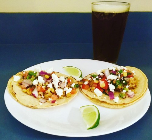 Two tacos topped with vegetables and cheese on a plate, accompanied by a glass of dark beverage and lime wedges.