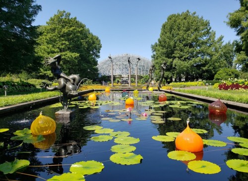 A serene garden scene featuring sculptures, colorful floating pumpkins, and lily pads in a tranquil pond.