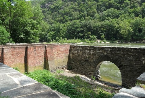 A stone arch bridge with a brick wall beside a river, surrounded by lush green trees and hills.
