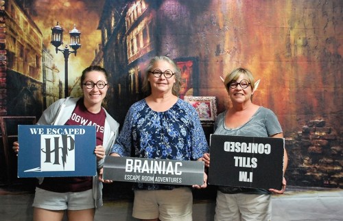 Three women hold signs celebrating their escape room experience, smiling in front of a themed backdrop.