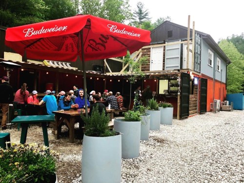 A lively outdoor gathering at a rustic venue with a Budweiser umbrella, surrounded by greenery and shipping container architecture.