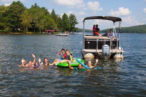 A group of people swimming and playing in a lake, with a boat nearby and trees in the background on a sunny day.