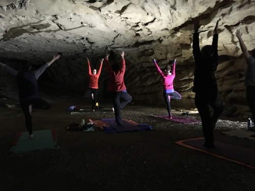 A group of people practicing yoga in a cave, with some in tree pose and others in various positions.