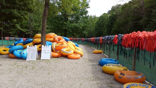 A pile of colorful inflatable tubes beside a row of life jackets on display in a wooded area.