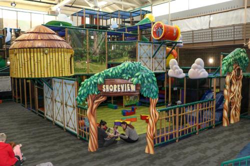 Colorful indoor playground with climbing structures, slides, and a treehouse theme, featuring children playing nearby.