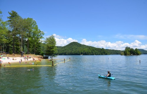 A serene lake scene with people swimming and a kayaker, surrounded by lush green trees and mountains under a clear blue sky.