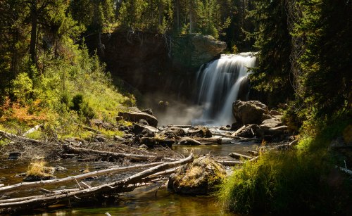 A serene waterfall cascades over rocks, surrounded by lush greenery and trees, with mist rising in the sunlight.