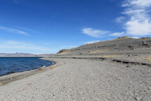 A rocky shoreline curves along a calm blue lake under a clear sky with scattered clouds.