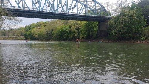 A serene river scene with kayakers paddling under a bridge, surrounded by lush greenery and calm waters.
