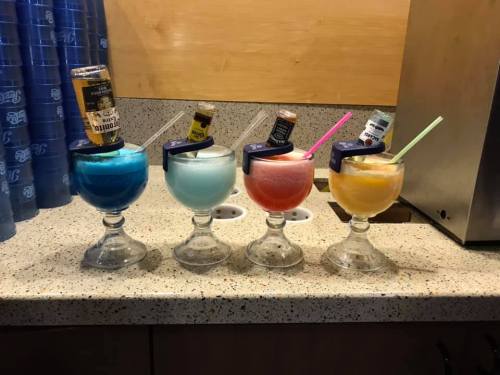 Four colorful frozen cocktails in glasses, each topped with a mini bottle and a straw, arranged on a countertop.