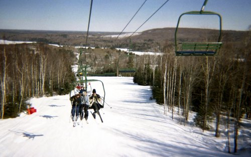 Skiers on a chairlift ascend a snowy slope, surrounded by trees and a scenic winter landscape.