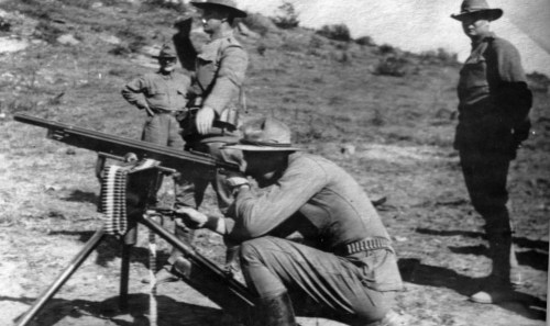 Black and white photo of soldiers operating a machine gun in a field, with two others observing in the background.