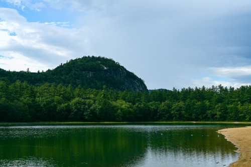 A serene lake surrounded by lush green trees and a distant hill under a partly cloudy sky.