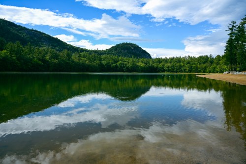 A serene lake reflecting blue skies and green trees, surrounded by mountains under a partly cloudy sky.