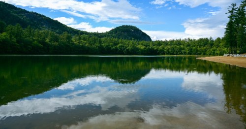 A serene lake reflecting trees and a mountain under a blue sky with scattered clouds.
