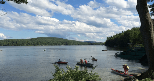 A serene lake scene with people kayaking, surrounded by green hills and a blue sky with fluffy clouds.