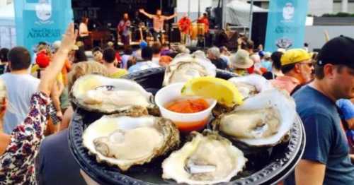 A platter of oysters with a lemon wedge and cocktail sauce, set against a lively outdoor festival crowd.