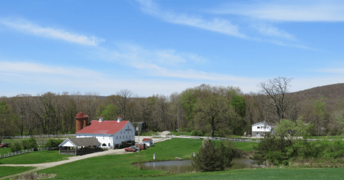 A scenic farm with a red-roofed barn, white buildings, trees, and a pond under a blue sky with wispy clouds.