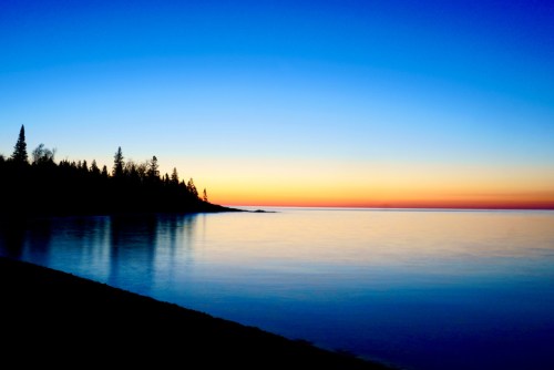 A serene sunset over calm waters, with silhouetted trees along the shoreline against a gradient sky.