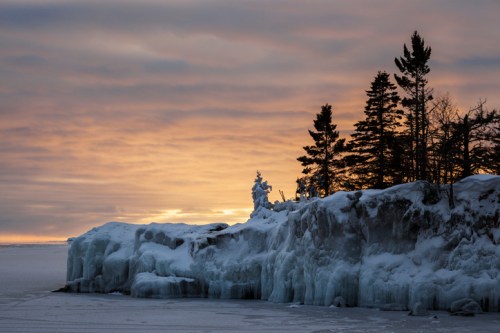 A snowy landscape with icy cliffs and evergreen trees silhouetted against a colorful sunset sky.