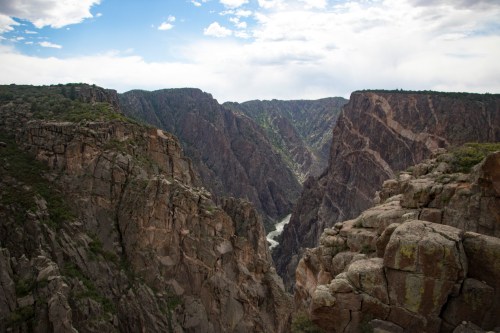 A dramatic canyon landscape with steep rocky cliffs and a winding river below, under a partly cloudy sky.