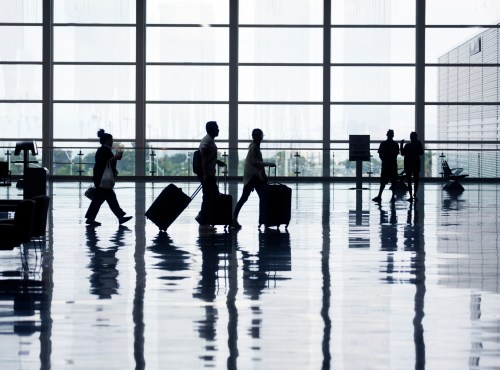 Silhouettes of travelers with luggage walking through a modern airport terminal with large windows.