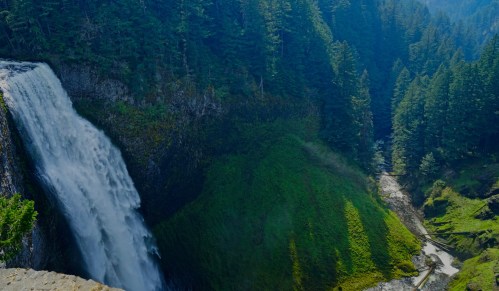 A stunning waterfall cascades down rocky cliffs, surrounded by lush green trees and a winding river below.