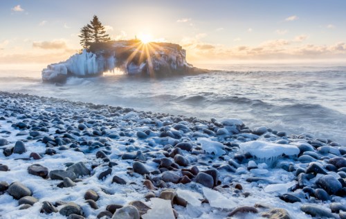 A rocky beach with ice and snow, featuring a small island with a tree, illuminated by a sunrise in the background.