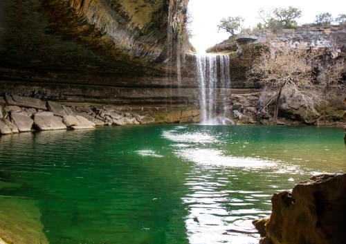 A serene waterfall cascades into a turquoise pool surrounded by rocky cliffs and sparse trees.