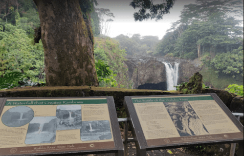 Two informational signs in front of a lush landscape featuring a waterfall, surrounded by greenery and mist.