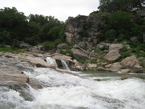 A rocky riverbank with cascading water and lush greenery in the background under a cloudy sky.