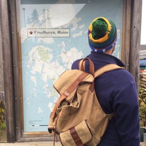 A person with a backpack examines a map of Vinalhaven, Maine, outdoors near a wooden structure.