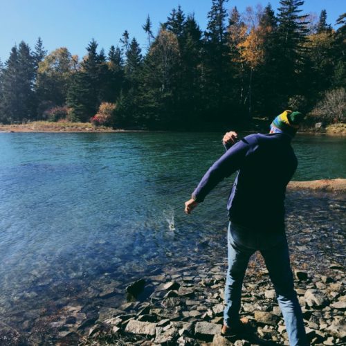 A person in a blue jacket and colorful hat stands by a serene lake, surrounded by trees in autumn colors.