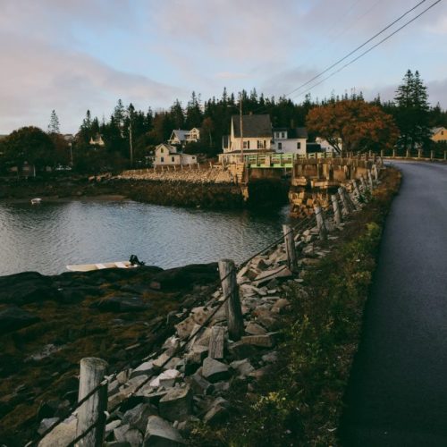 A scenic view of a coastal road with houses, trees, and a calm waterway lined with rocks and a small boat.