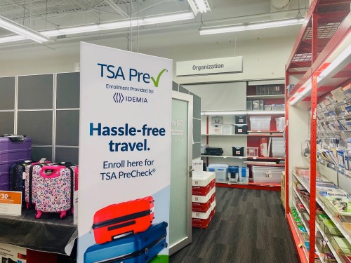 A sign for TSA PreCheck enrollment in a store aisle with luggage and organizational supplies in the background.