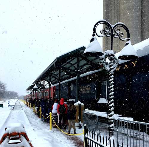 A snowy scene with people waiting in line at a train station, surrounded by snow-covered buildings and a lamppost.