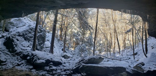 A snowy landscape viewed through a rocky cave opening, with trees and sunlight filtering through.