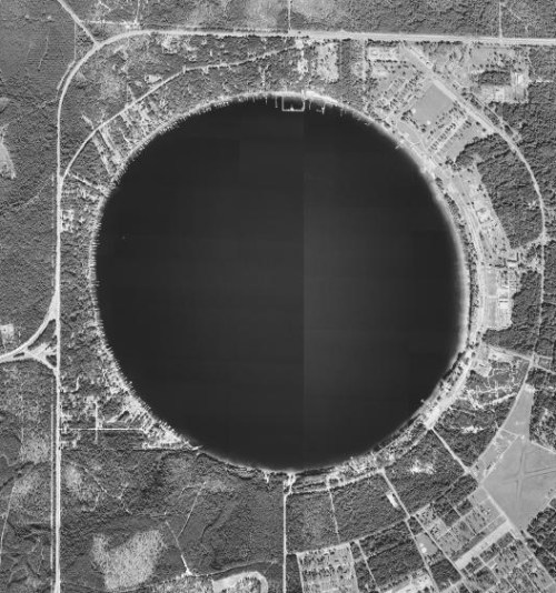 Aerial view of a large, circular lake surrounded by trees and roads, with nearby residential areas visible.