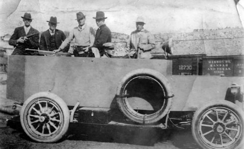 Five men in hats pose with an early automobile, showcasing vintage attire and a historical setting.