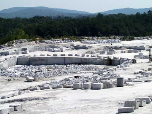 A large marble quarry with white stone blocks and machinery, surrounded by green trees and distant hills under a clear sky.