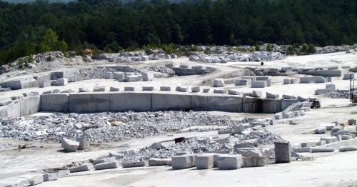 A large marble quarry with white stone blocks and a forested area in the background under a clear blue sky.