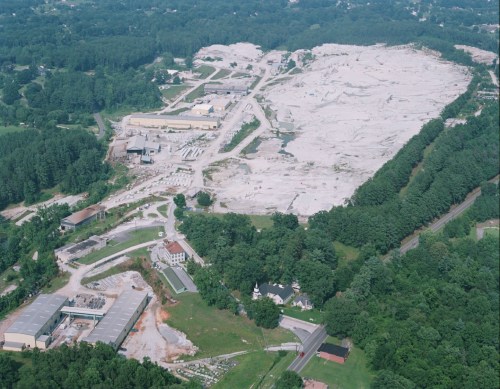 Aerial view of a large, cleared construction site surrounded by trees and buildings, with a gravel surface and machinery visible.