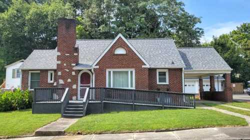 A brick house with a front porch, large windows, and a sloped roof, surrounded by greenery and a sidewalk.