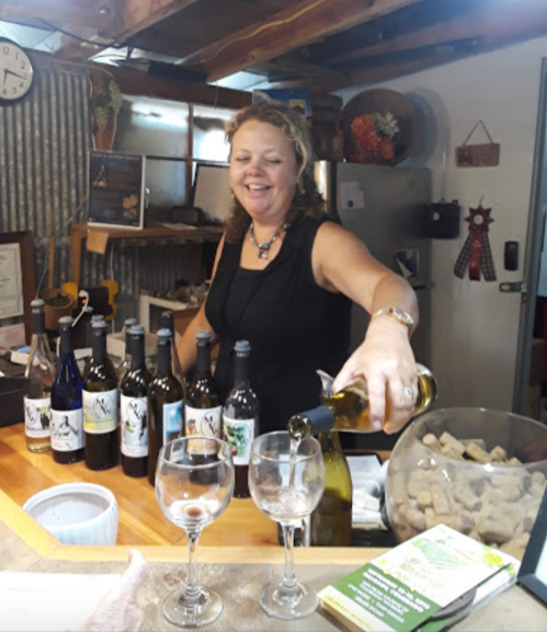 A woman pours wine at a rustic bar, surrounded by bottles and glasses, smiling warmly.