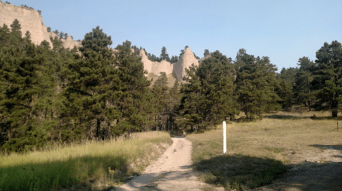 A dirt path winds through a forested area with tall trees and rocky cliffs in the background under a clear blue sky.